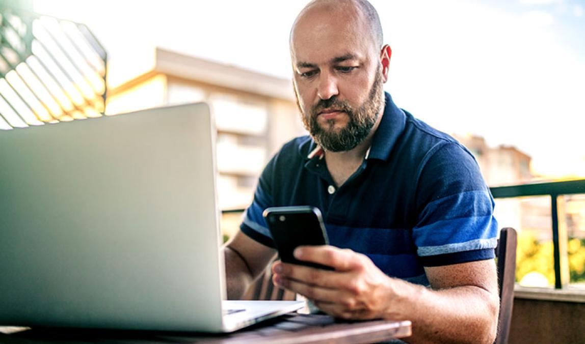 Bearded male sitting at outdoor table using laptop looking at smart mobile device in left hand