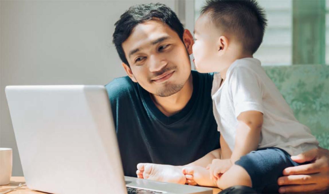 A man smiling looking at his computer with a toddler.